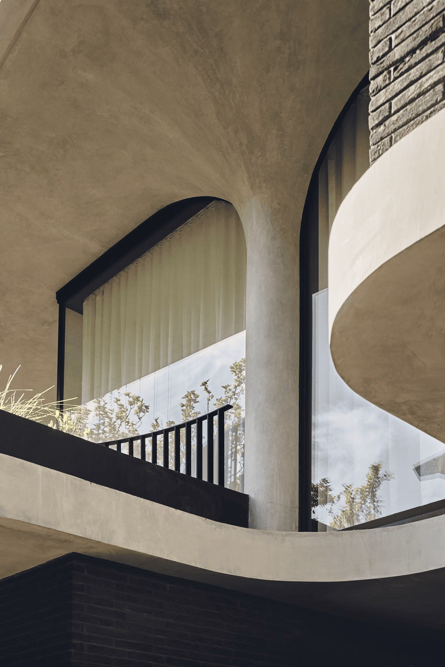 Curved modern concrete balcony with glass windows and vertical black railing reflecting nearby trees.