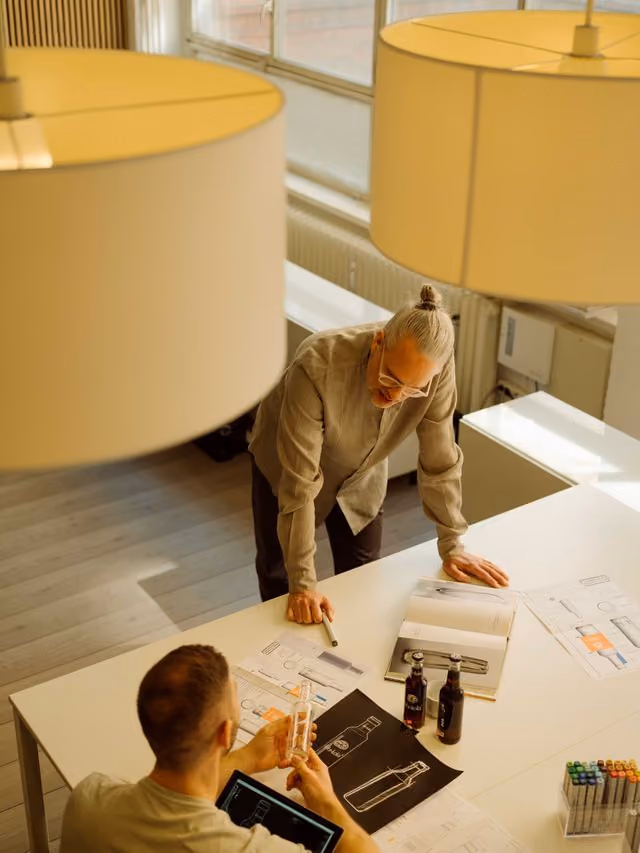 Two men reviewing bottle design sketches and prototypes at a white table in a well-lit room.