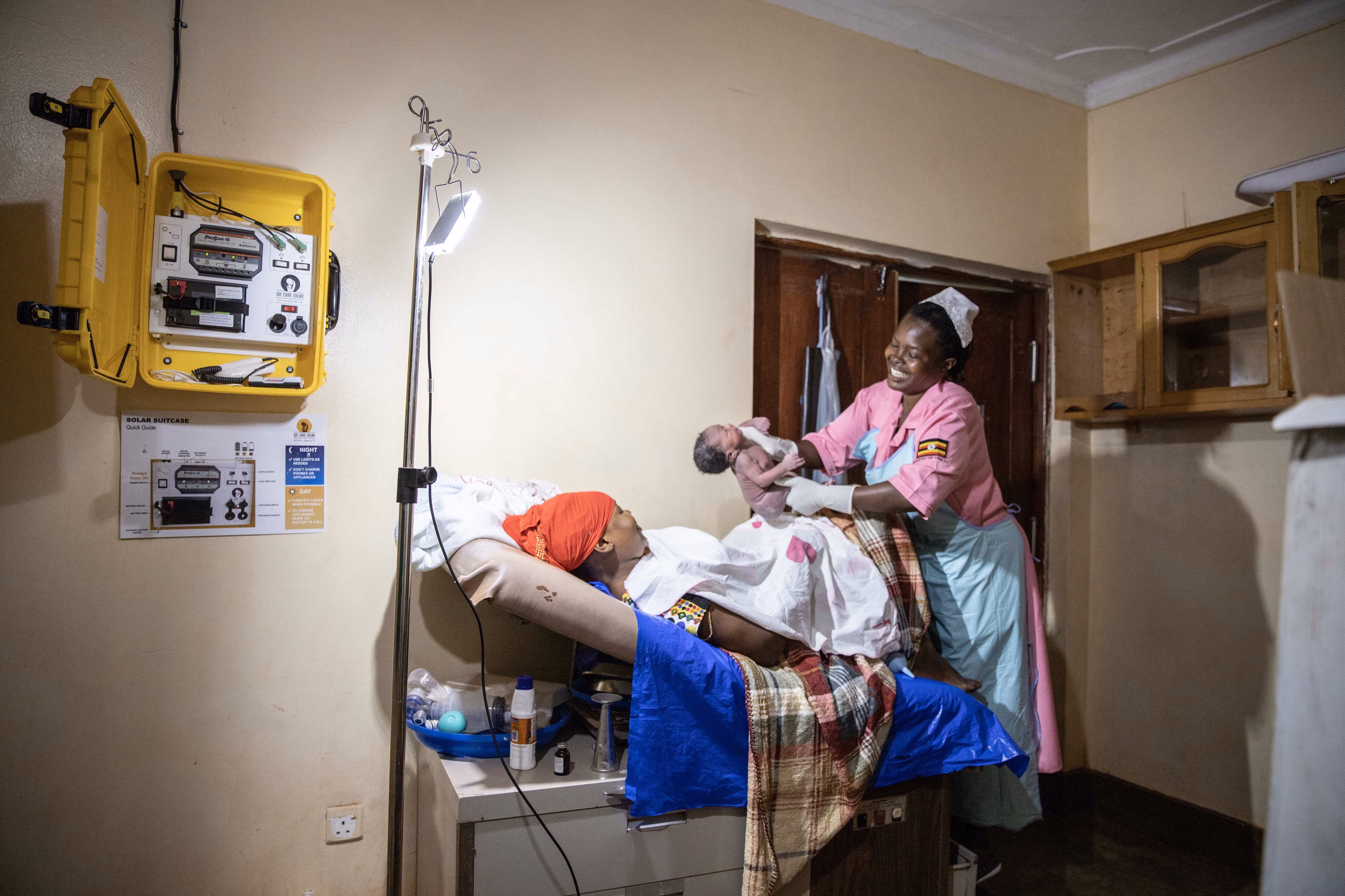 Nurse in pink uniform holding newborn baby while mother lies on bed in a dimly lit room.