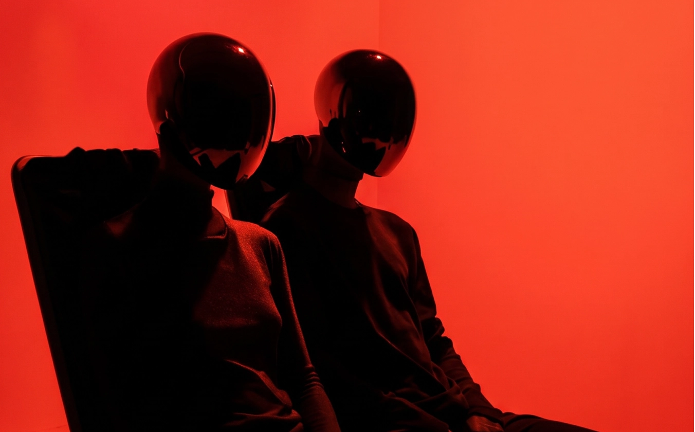 Two seated figures wearing black reflective helmets and dark clothes against a red background.
