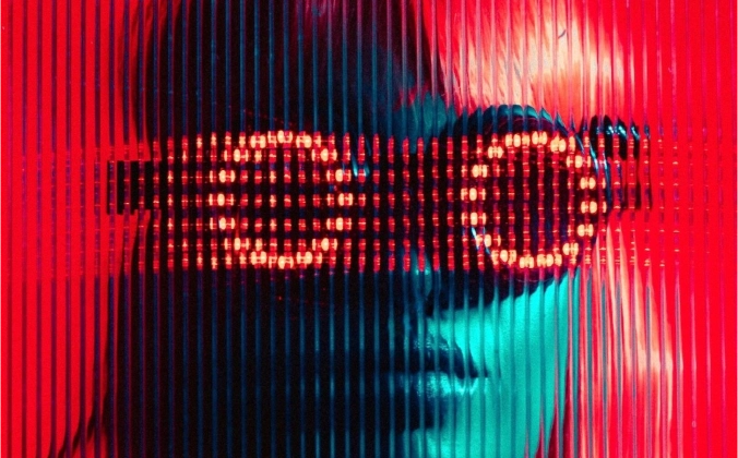 Person wearing LED light sunglasses with circular patterns behind a ribbed glass panel with red and blue lighting.