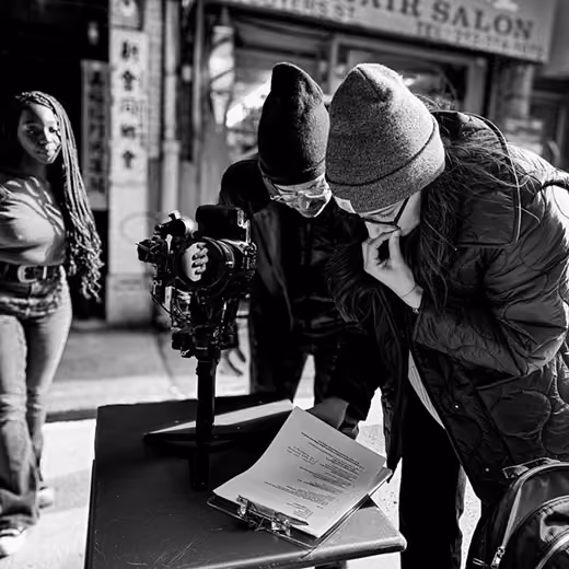Black and white photo of a crew reviewing a script beside a camera on a city street