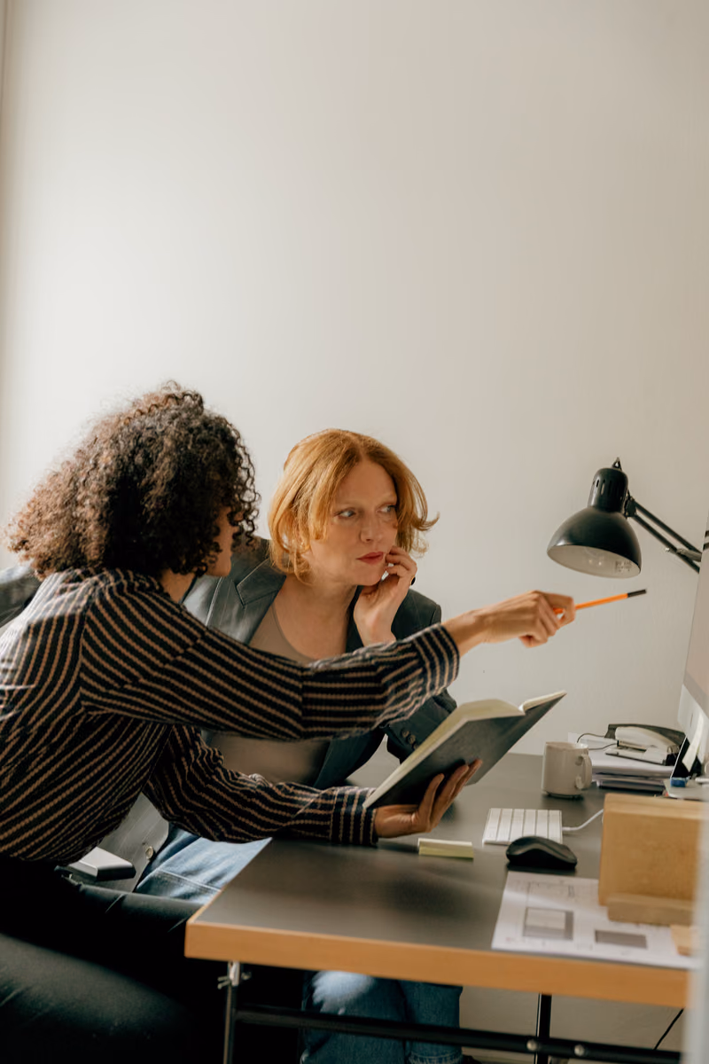 Two art directors reviewing a book at a desk, one pointing with a pencil