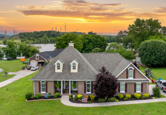 A large brick house with a shingled roof, front porch with plants, and well-maintained landscaping, set against a sunset sky and near a river with trees in the background.