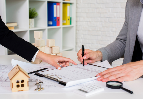 Two people at a desk reviewing and signing estate or legal documents, surrounded by a small wooden house model, keys, a calculator, magnifying glass, and building blocks on a white table.