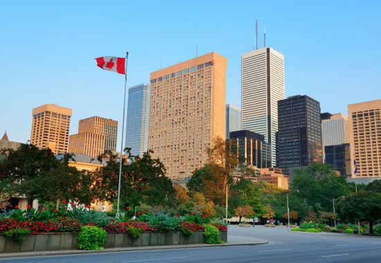 Cityscape with tall modern buildings behind lush green trees and colorful flower beds, featuring a Canadian flag on a flagpole under a clear blue sky.