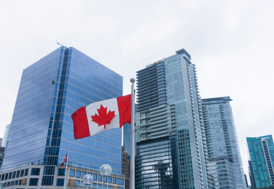 Canadian flag flying on a pole with modern glass skyscrapers in the background under a cloudy sky.