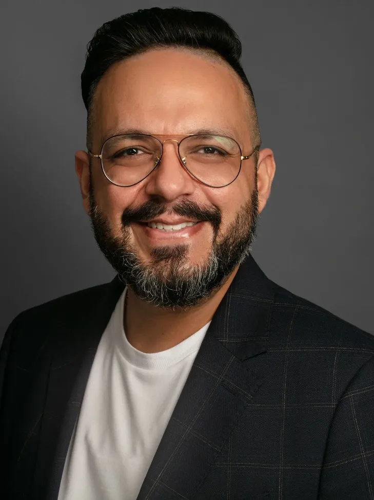 Smiling man with a neatly trimmed beard and mustache wearing round glasses, a white shirt, and a dark blazer against a gray background.