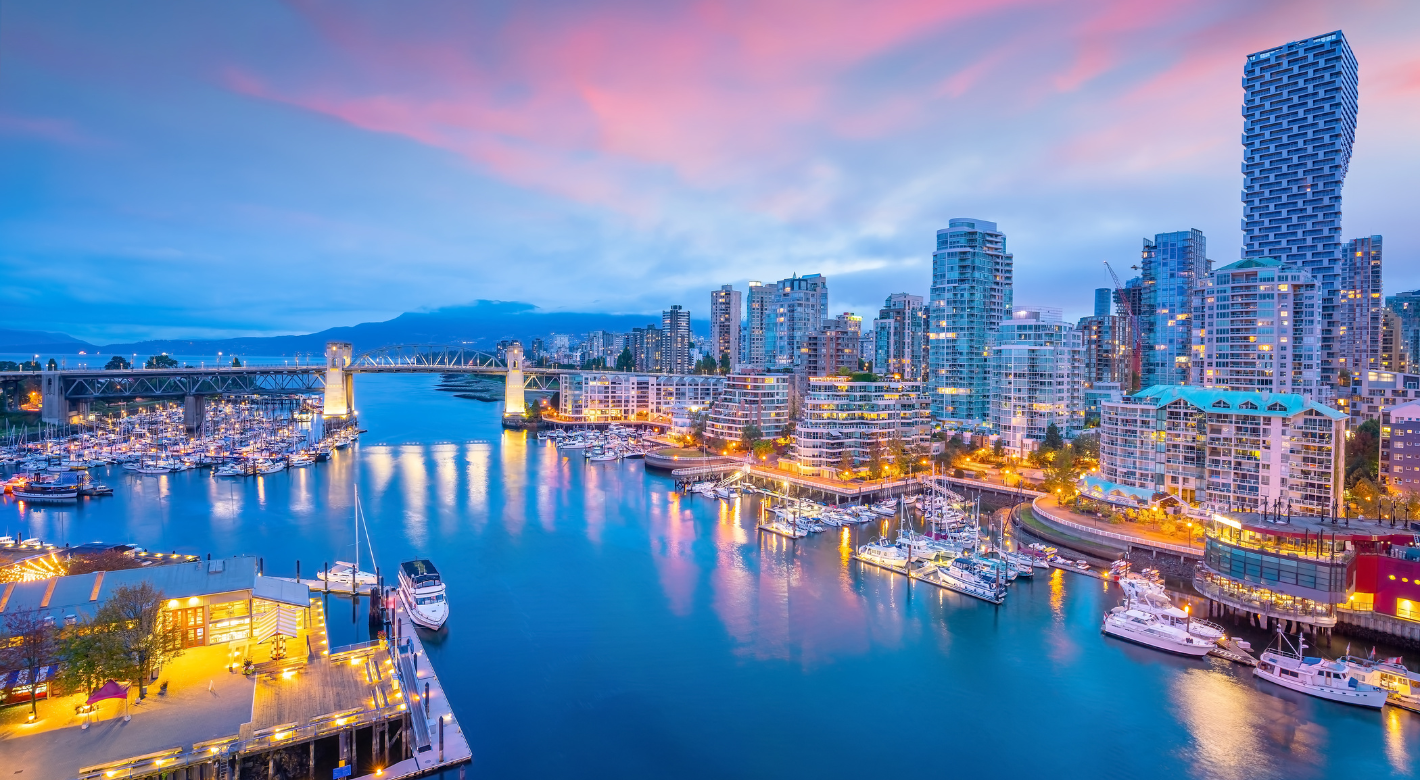 Evening skyline of a coastal city with illuminated high-rise buildings, a marina full of boats, a lit waterfront, and a bridge spanning a calm body of water under a pink and blue sky.
