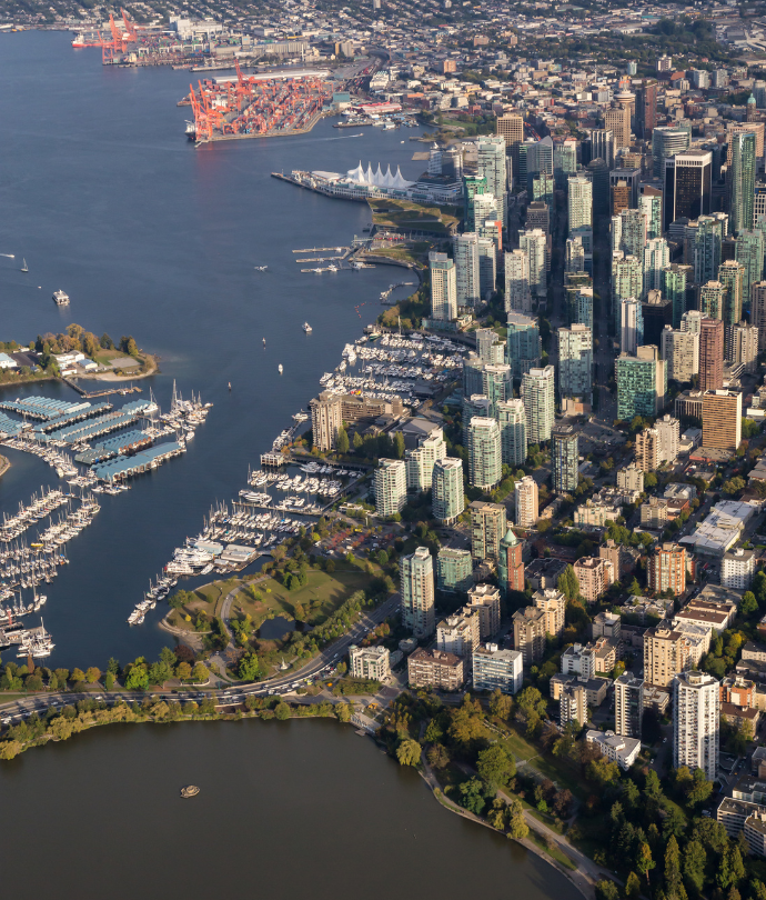 Aerial view of a coastal cityscape featuring dense high-rise buildings, a harbor with numerous boats and yachts docked, marina, industrial cranes, and waterfront parks with green spaces.