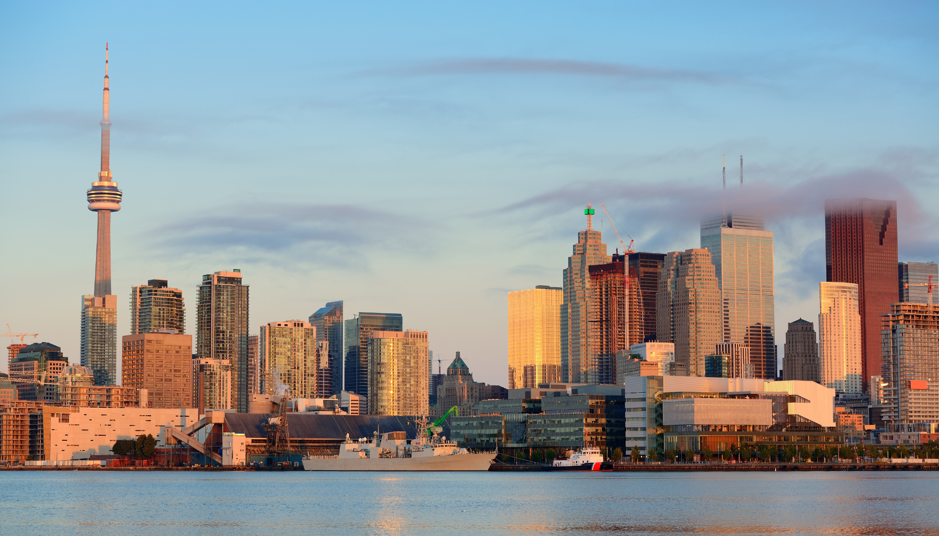 Toronto skyline at sunset featuring the CN Tower, high-rise buildings with sunlight reflections, light clouds partially covering some skyscrapers, and ships docked along the waterfront.