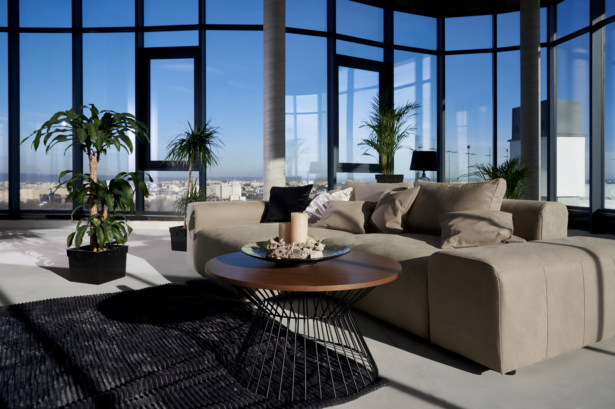 Modern living room with a large beige sectional sofa adorned with cushions, a round wooden coffee table with a decorative centerpiece of candles and stones, black textured rug, several potted green plants, and floor-to-ceiling windows offering a panoramic city view under a clear blue sky.
