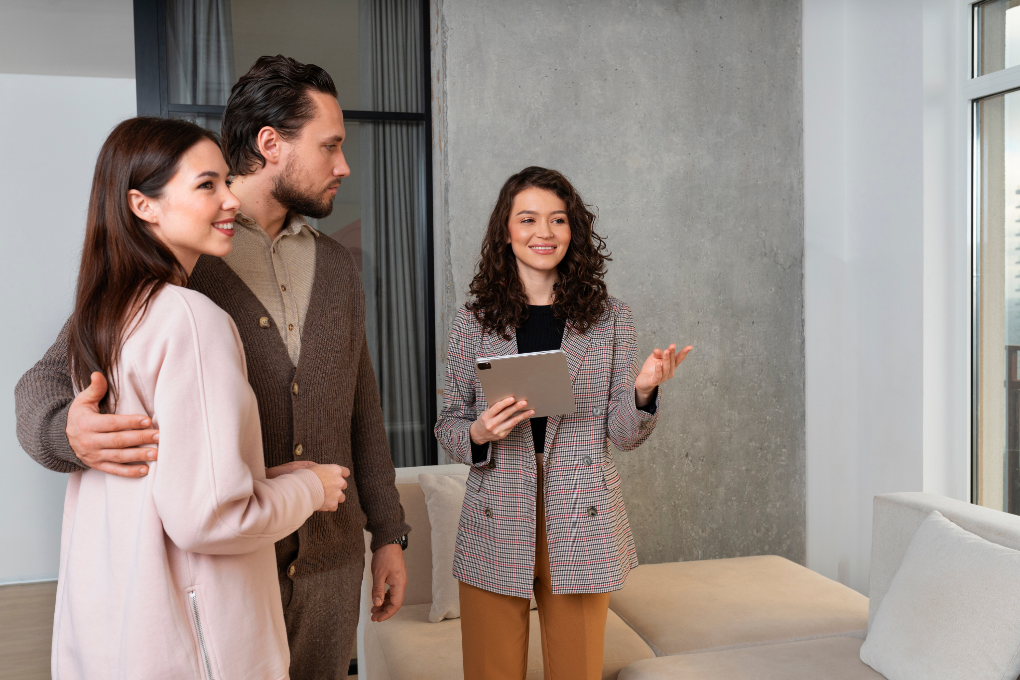 A real estate agent holding a tablet shows a couple a modern apartment interior featuring a beige sectional sofa and a large window.