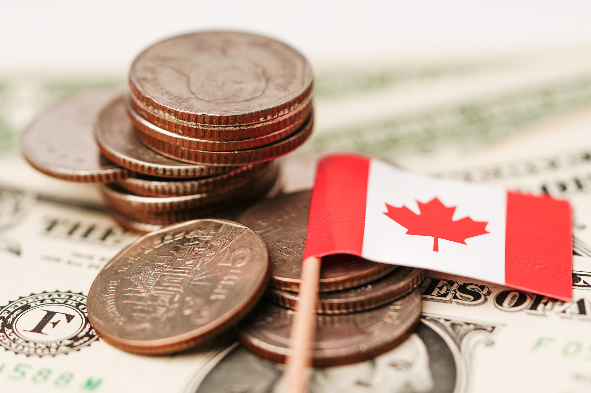 Stack of coins and a small Canadian flag on top of a U.S. dollar bill, symbolizing currency and economic relations.