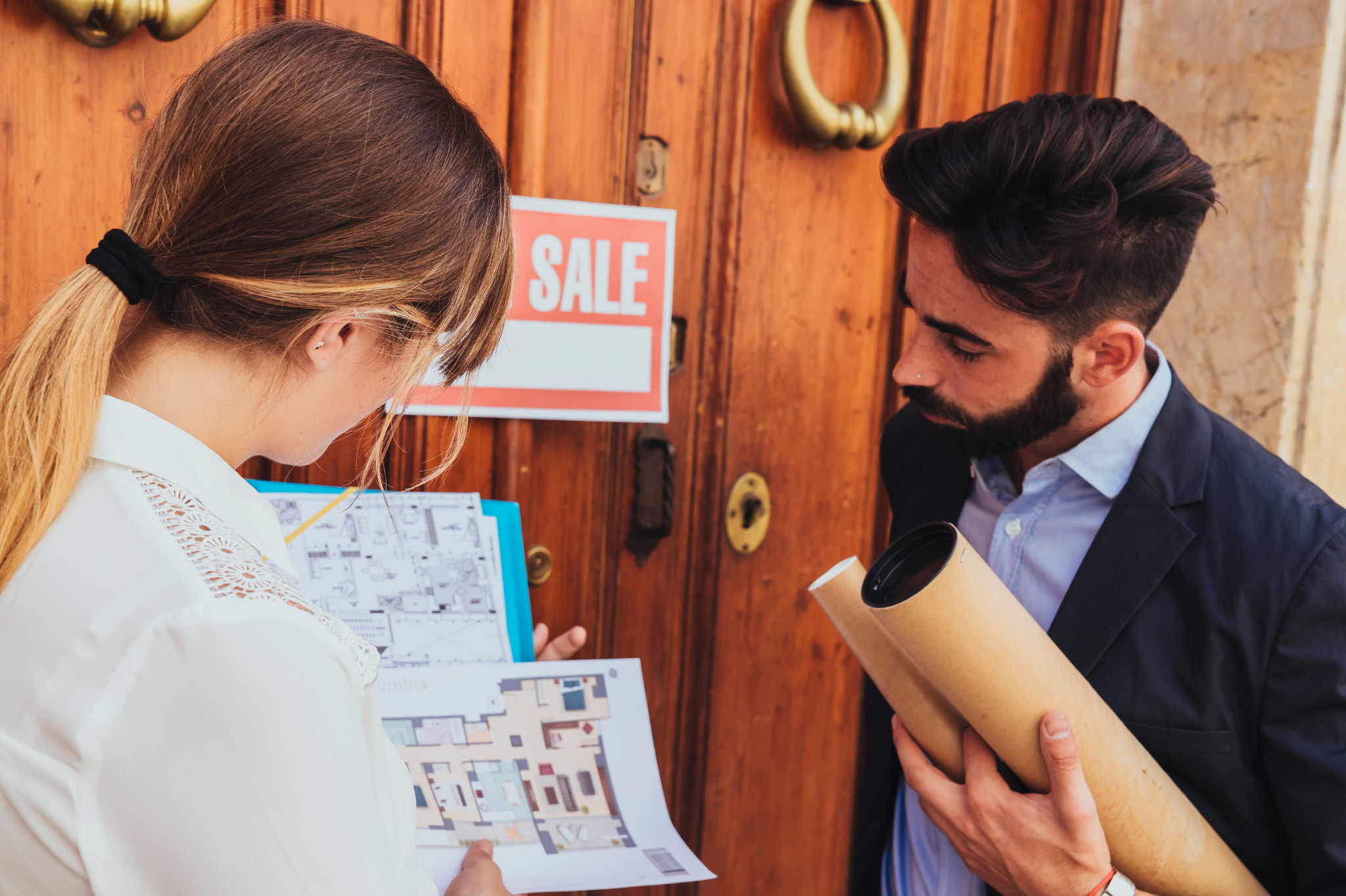 Two people, a woman and a man, are standing in front of a wooden door with a 'SALE' sign. The woman holds floor plans on a clipboard, and the man, dressed in a blazer, holds rolled-up architectural plans, both looking at the documents.