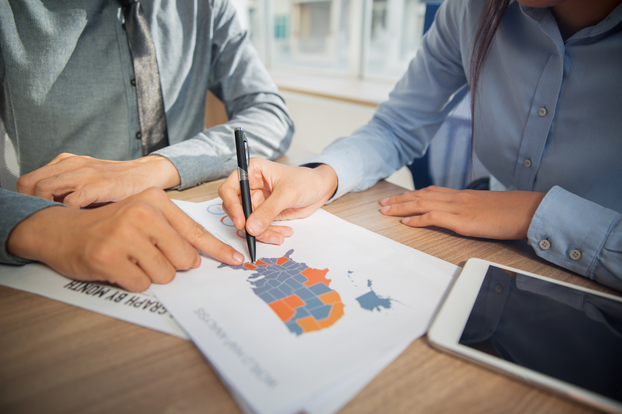 Two people in business attire reviewing a color-coded map on paper at a desk, one pointing and the other marking with a pen, with a tablet nearby.