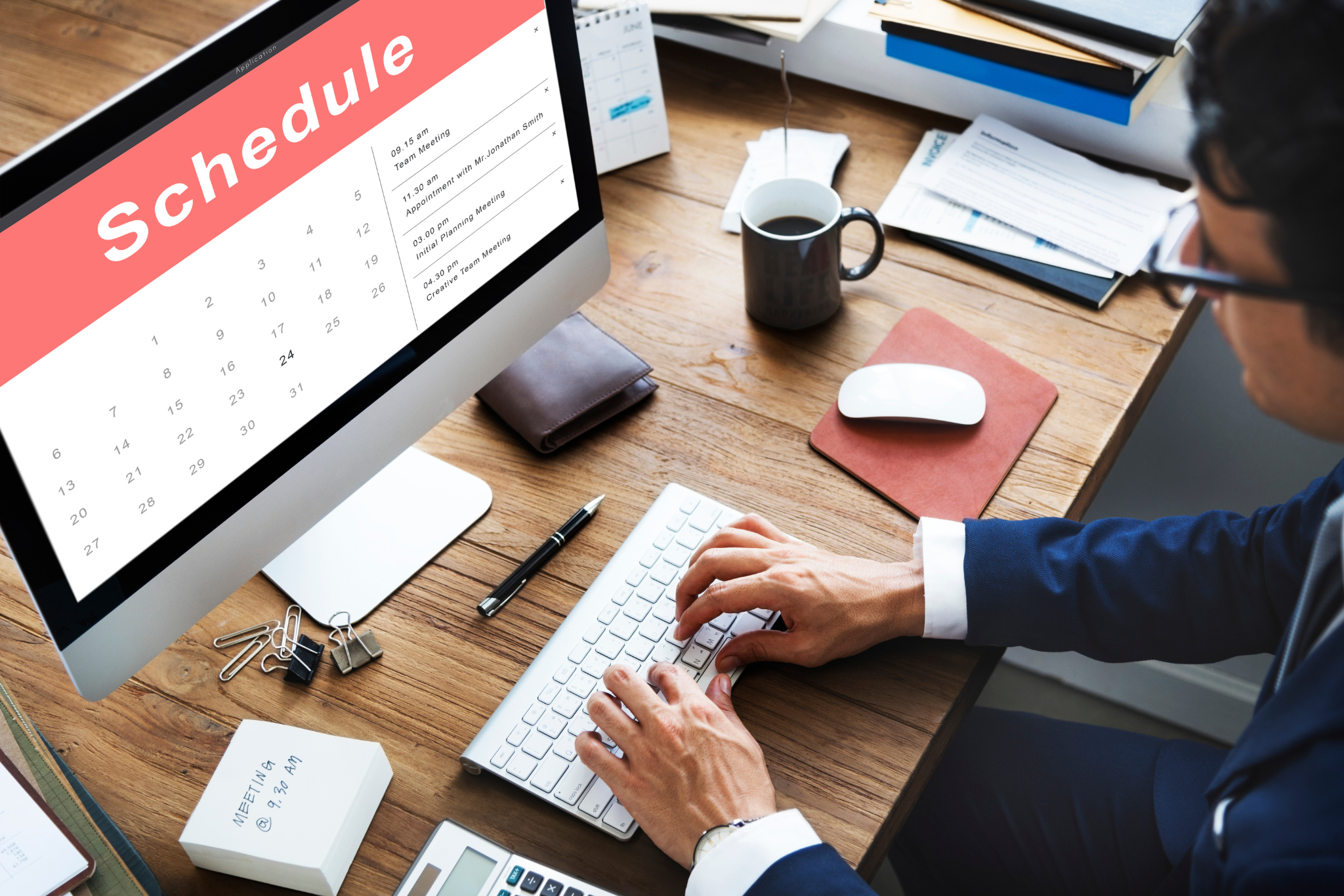 Person in a business suit typing on a white keyboard at a wooden desk with a coffee mug, papers, calculators, and a monitor displaying a schedule and calendar for June with meetings and appointments listed.