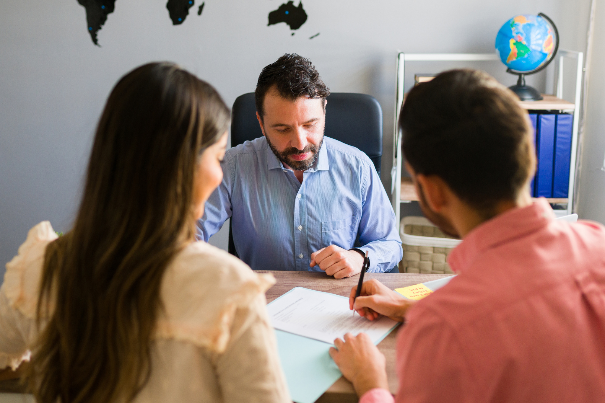 Man with dark hair and beard sitting behind a desk in an office, attentively watching a couple sign documents on the desk.