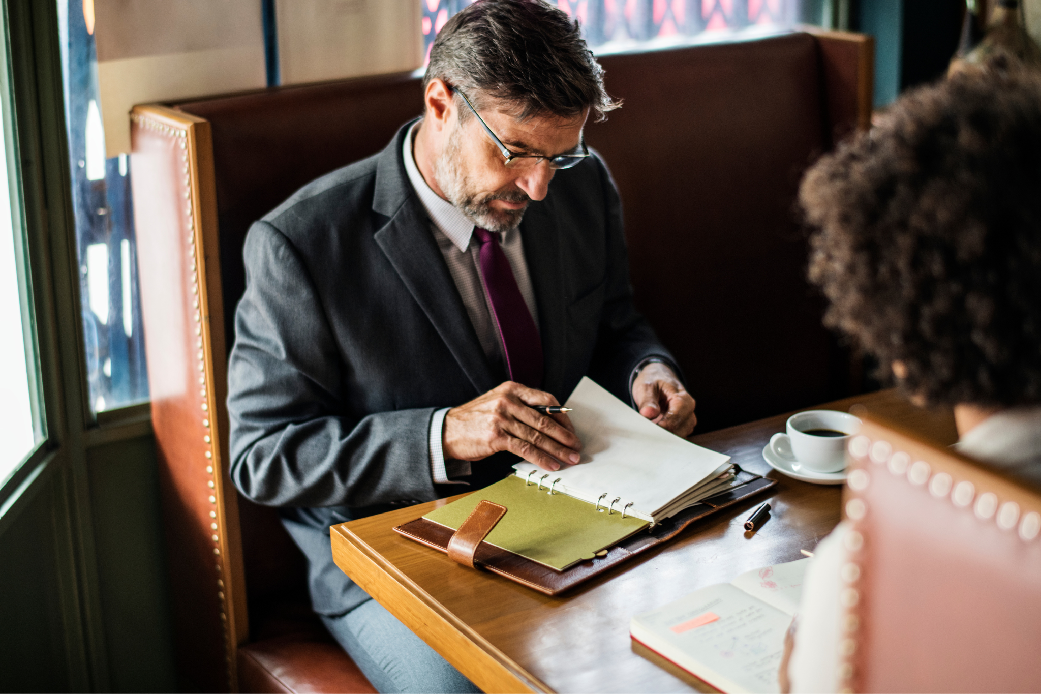 A man in a gray suit and glasses sitting at a wooden table reviewing papers in a binder opposite a person with curly hair who is holding a notebook, with a cup of coffee on the table.