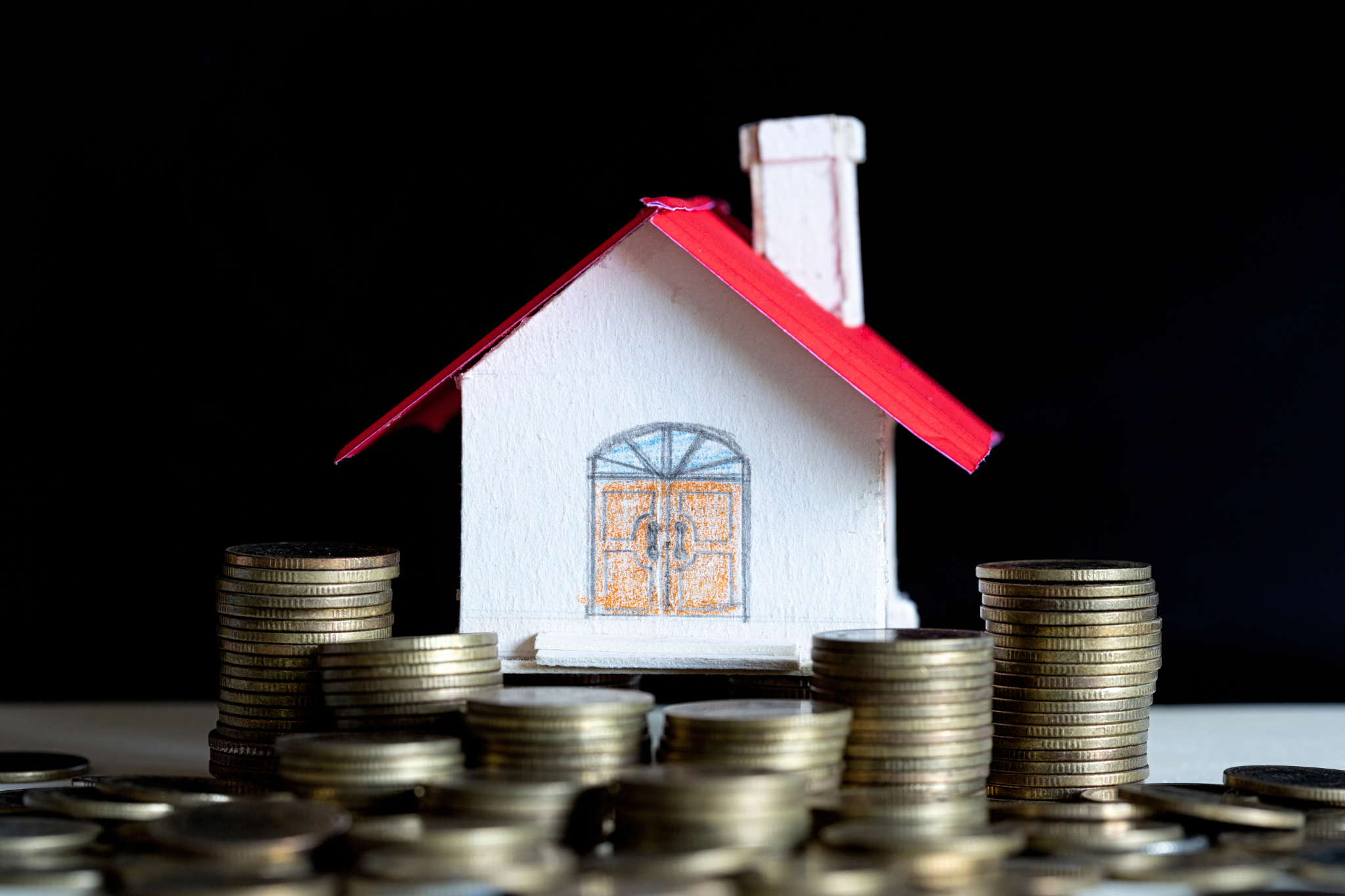 Small white house model with red roof surrounded by stacks of coins against a black background, symbolizing real estate investment or mortgage financing.