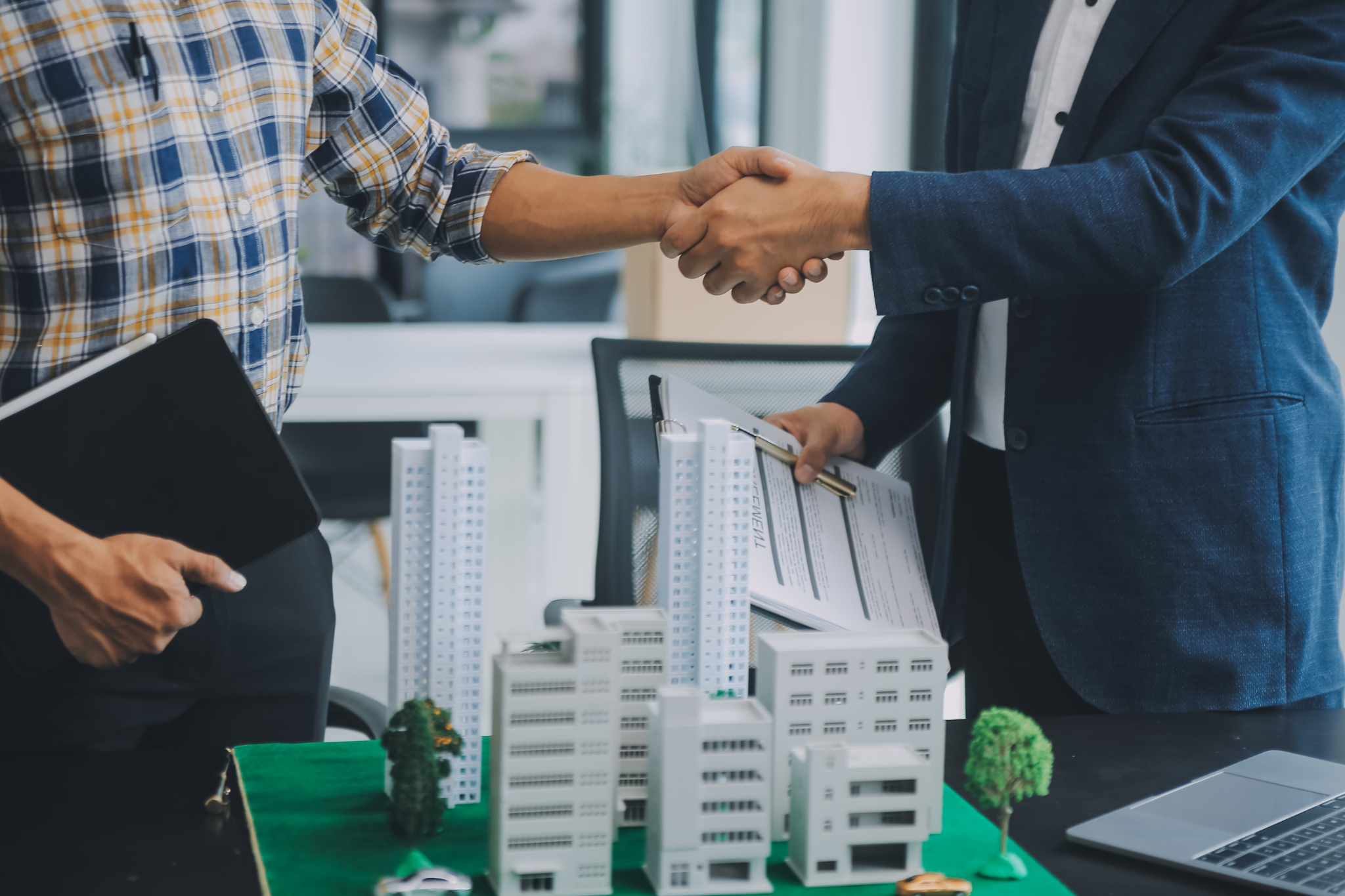 Two people shaking hands over a table with a model of buildings and trees, one holding a clipboard and the other holding a pen and documents, suggesting a business or real estate agreement.