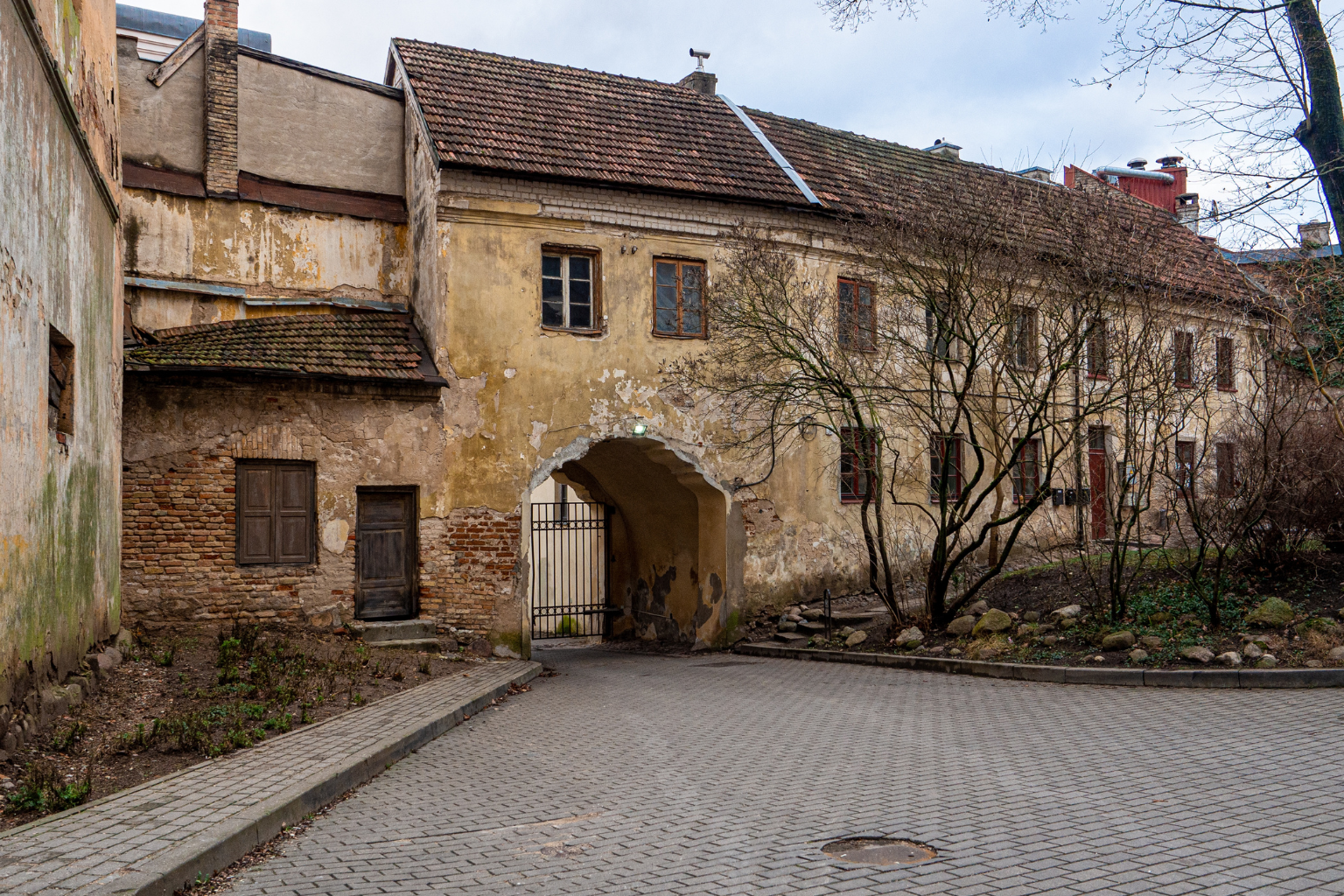 Old, weathered building with peeling yellowish plaster, wooden windows, and a tiled roof, featuring an arched passageway with a metal gate, surrounded by bare trees and a paved courtyard.