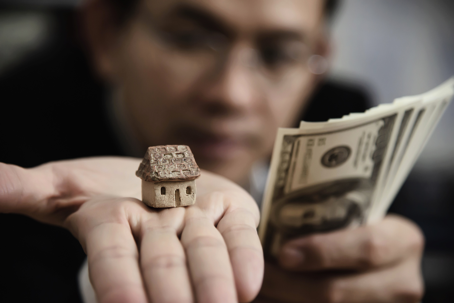 Person holding a small model house in one hand and fanning out several US hundred-dollar bills in the other, symbolizing real estate or mortgage finance.