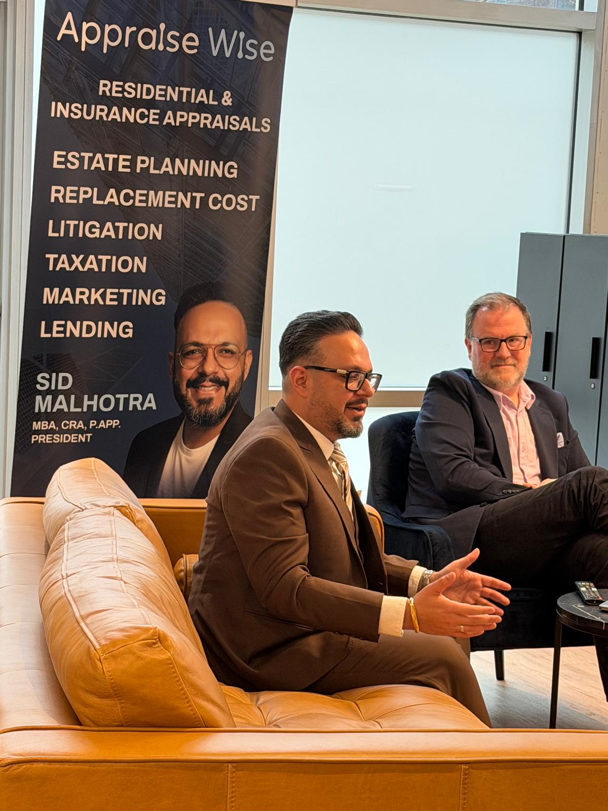 Two men in business attire seated in a modern office setting. One man in a brown suit gestures while speaking, the other in a dark blazer listens attentively. A large banner behind them displays the text 'Appraise Wise' and lists services including residential and insurance appraisals, estate planning, replacement cost, litigation, taxation, marketing, lending, with the name Sid Malhotra, President, MBA, CRA, P.APP.