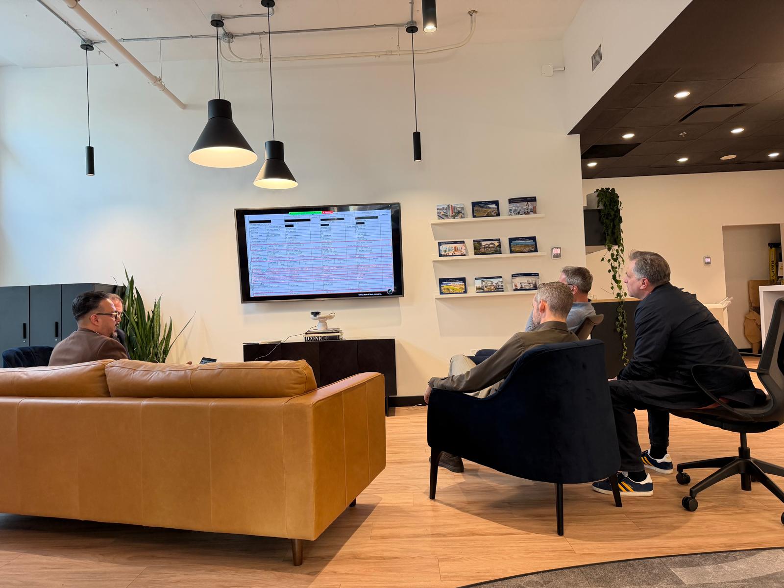 Four men in a modern office lounge area, sitting on a tan leather couch, a black armchair, and a black office chair, watching a wall-mounted flat screen displaying a spreadsheet with highlighted text. The room has hanging black pendant lights, plants, framed pictures on shelves, and a wooden floor.