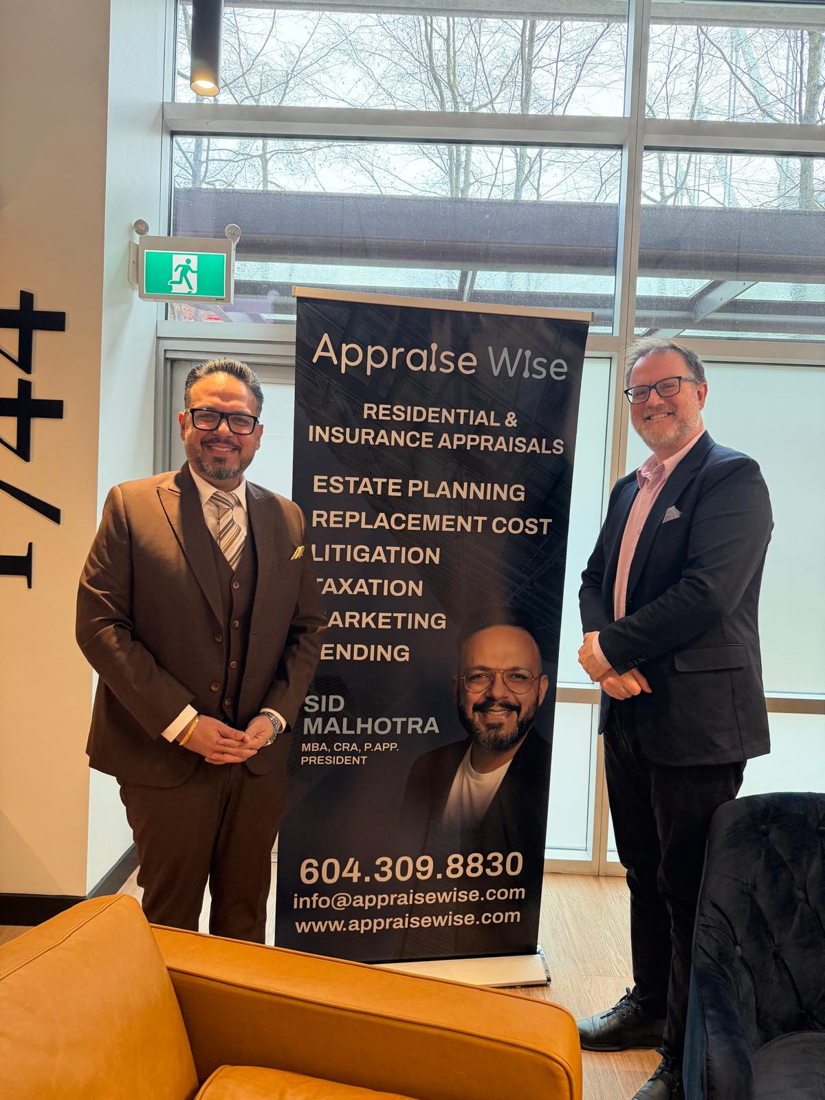 Two men in suits standing inside a modern office near a large window and a banner for Appraise Wise, listing services like residential and insurance appraisals, estate planning, litigation, and taxation, with contact details for Sid Malhotra, president.