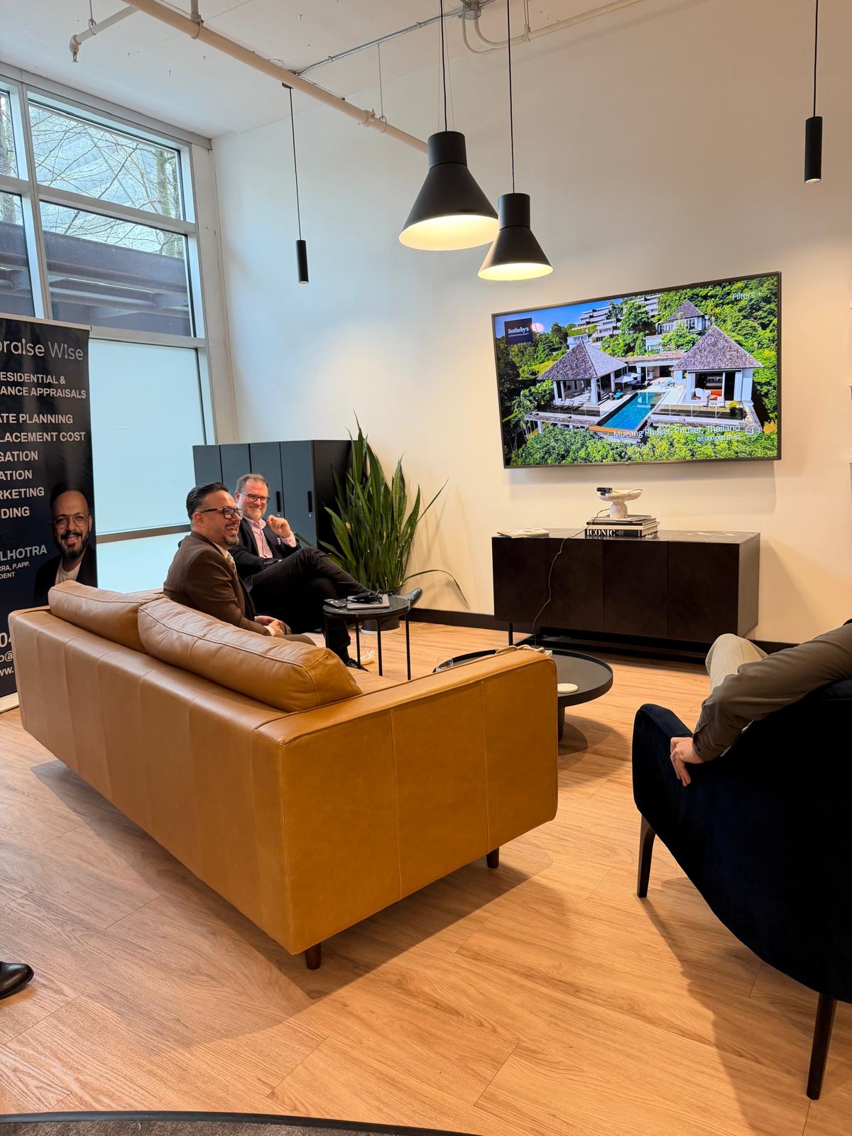 Three men sitting in a modern office lounge area with a brown leather couch and dark chairs, watching a large TV screen displaying an image of a luxurious waterfront property with a pool and thatched roofs.