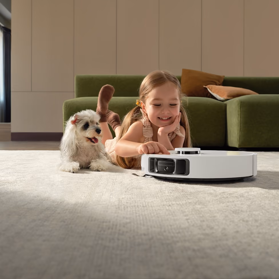 Smiling girl lying on carpet next to small white dog, interacting with a white robot vacuum cleaner.