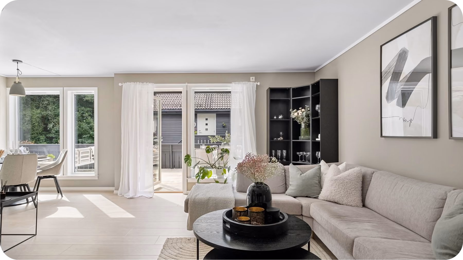 Bright living room with gray sectional sofa, black round coffee table, plants, and large windows with curtains.