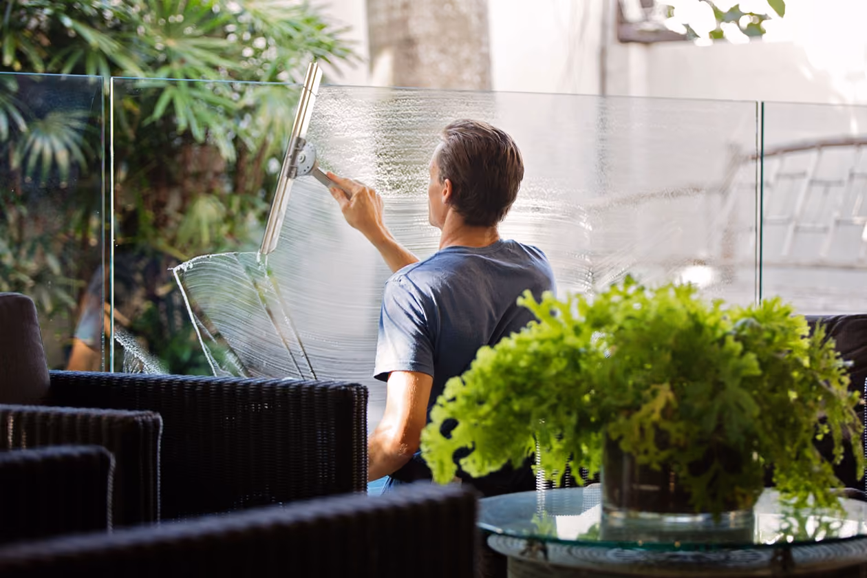 Man cleaning outdoor glass panel with a squeegee beside patio furniture and plants.