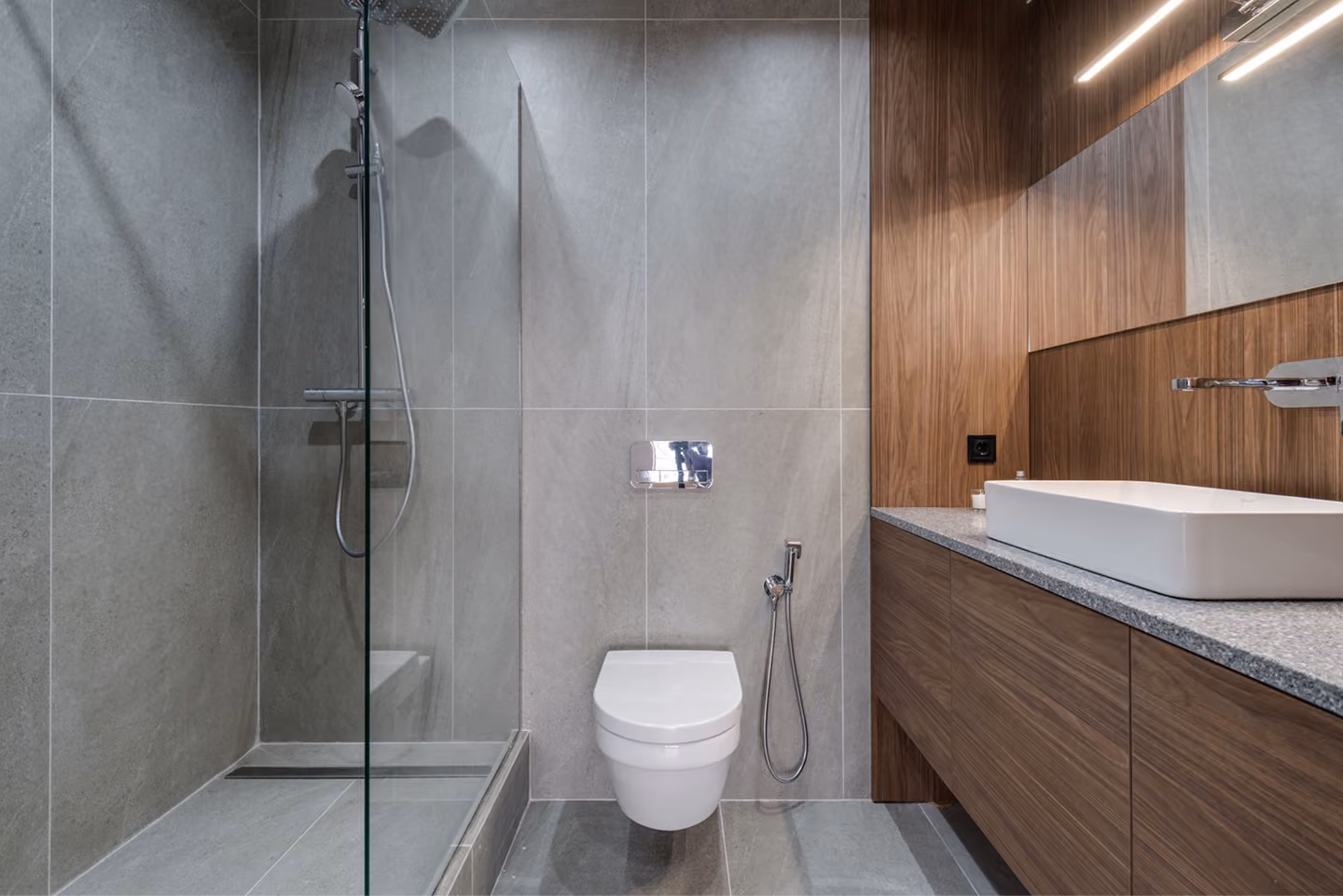 Modern bathroom with gray tile shower, wall-mounted toilet, and wooden vanity with rectangular sink.