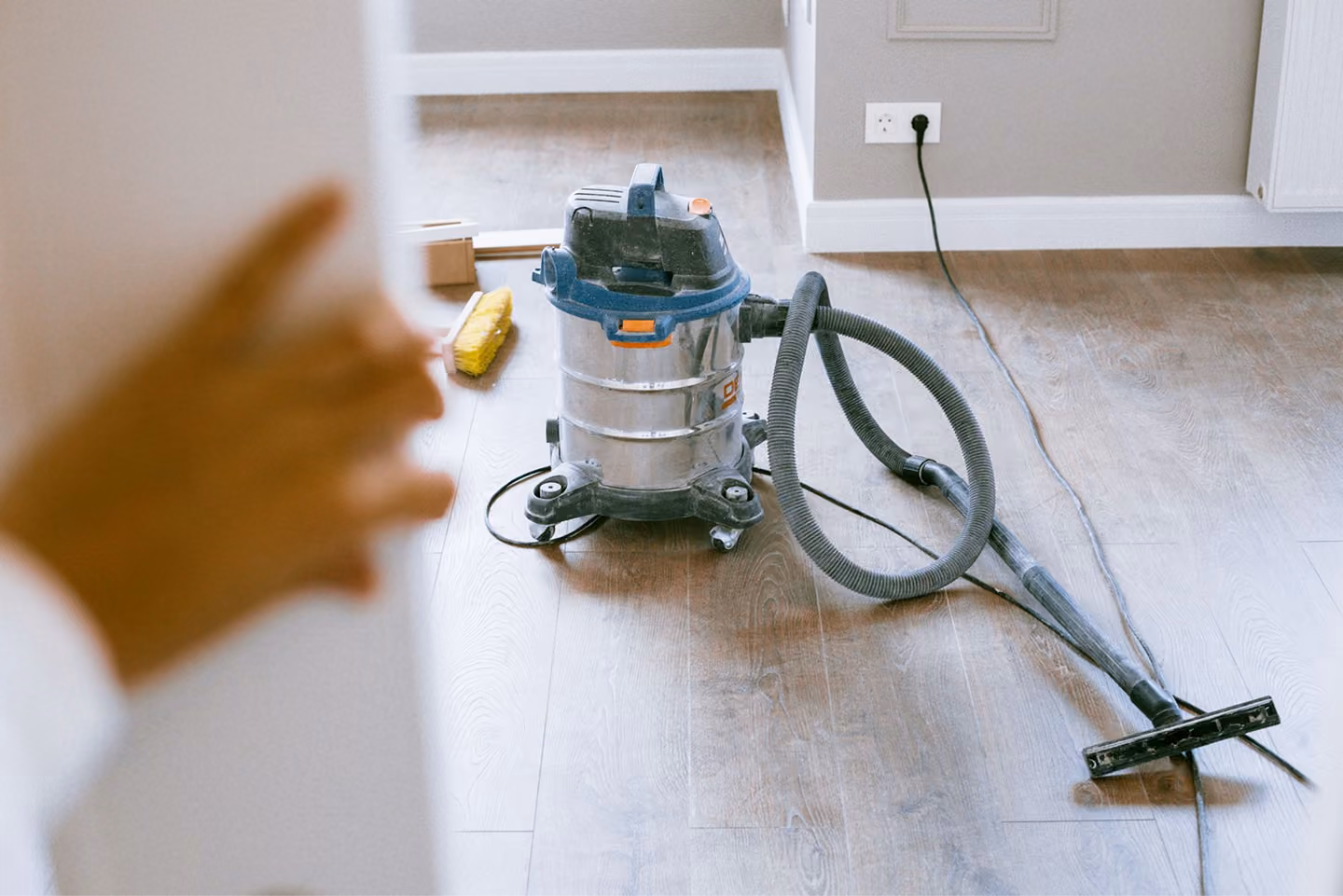 Vacuum cleaner on a wood floor near a wall outlet and a hand holding a door open.