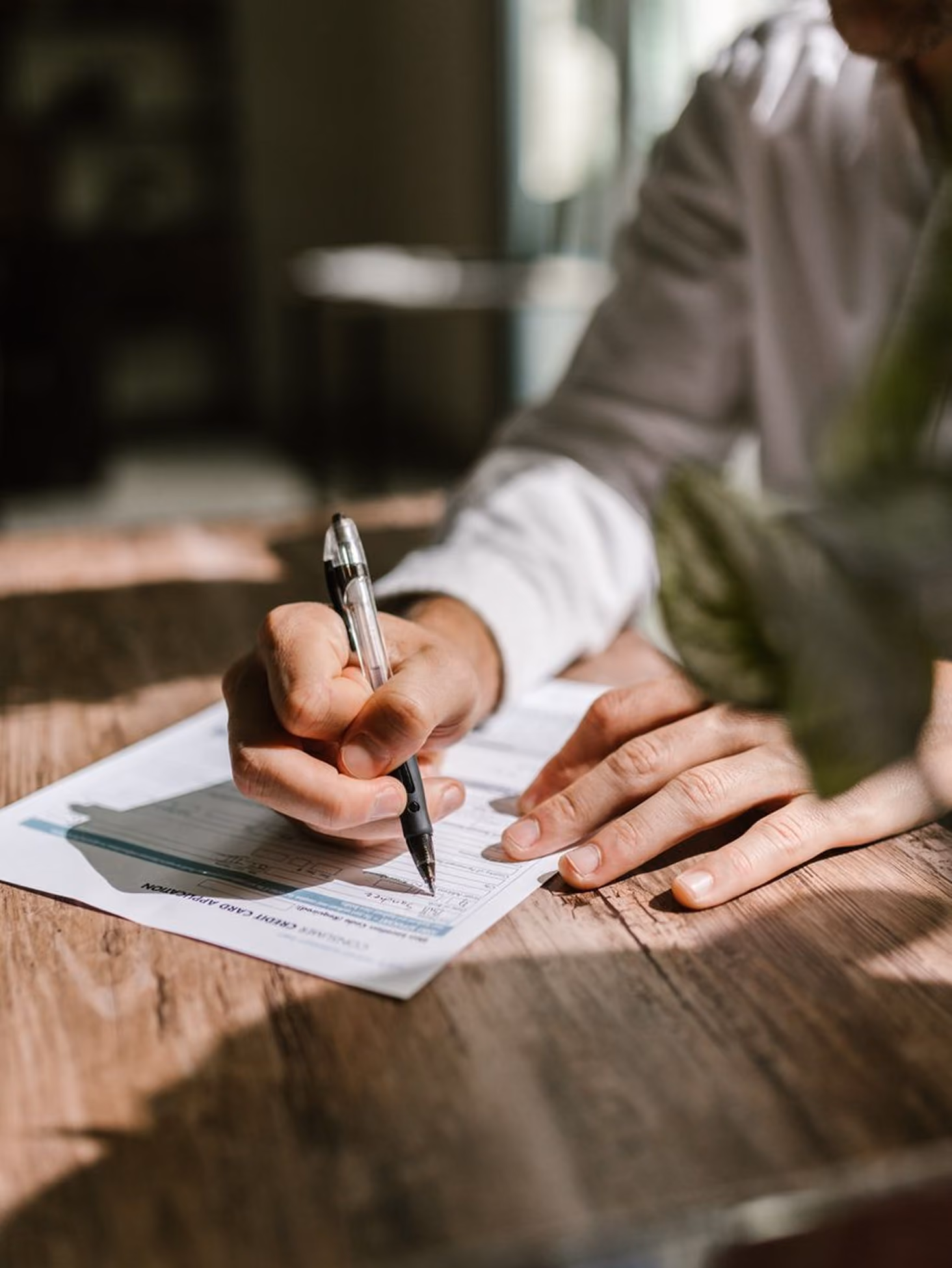 Person wearing white shirt filling out a credit card application form with a black pen on wooden table.