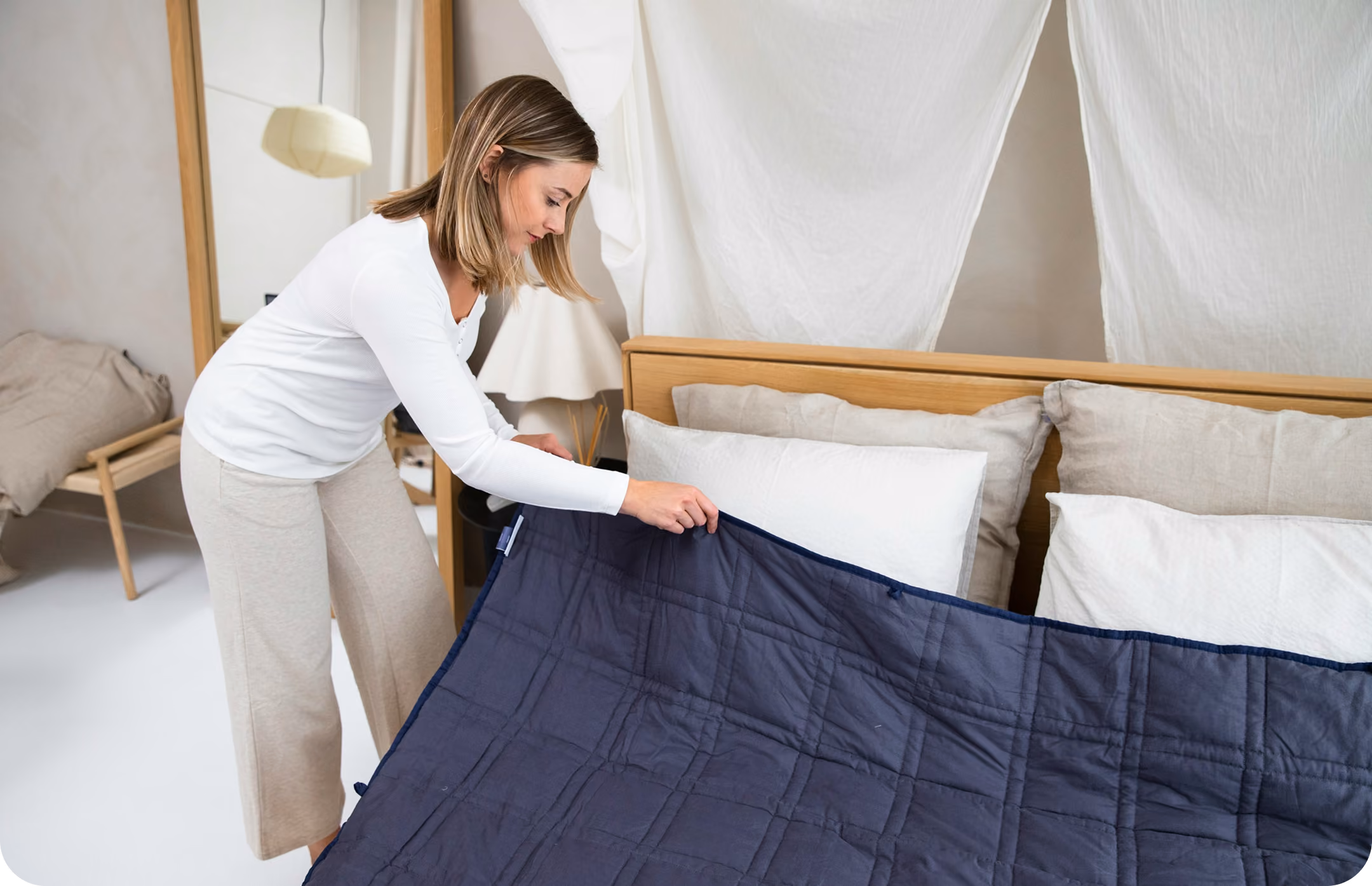 Woman in white shirt and beige pants spreads a dark blue quilt over a bed with white pillows.