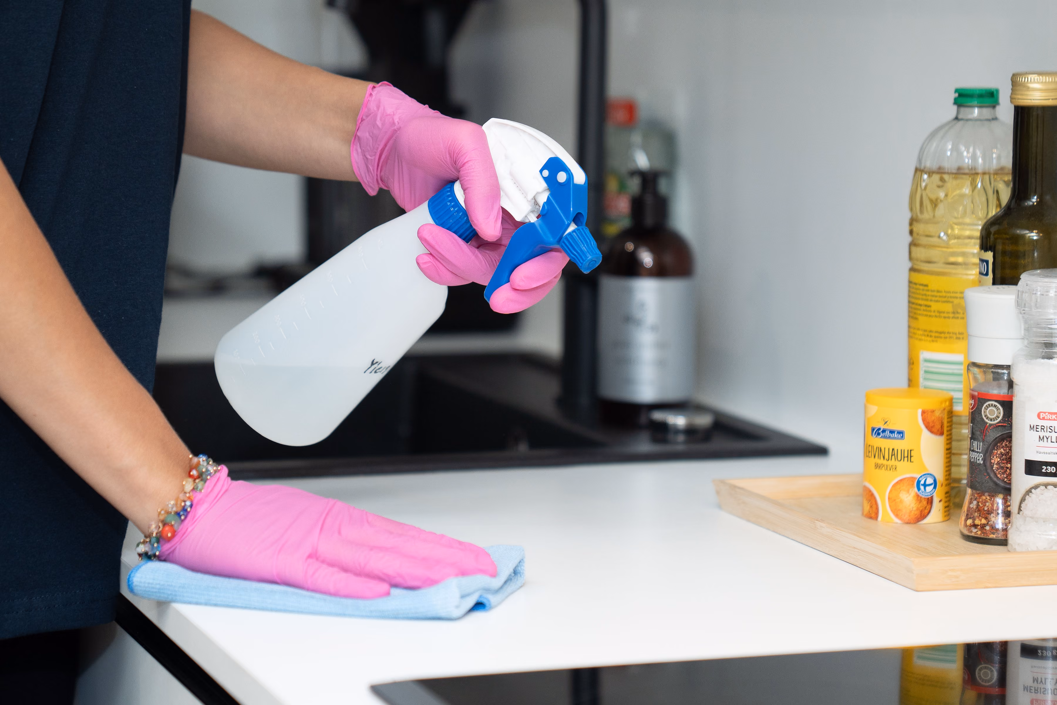 Person wearing pink gloves cleaning a white kitchen countertop with spray bottle and cloth.