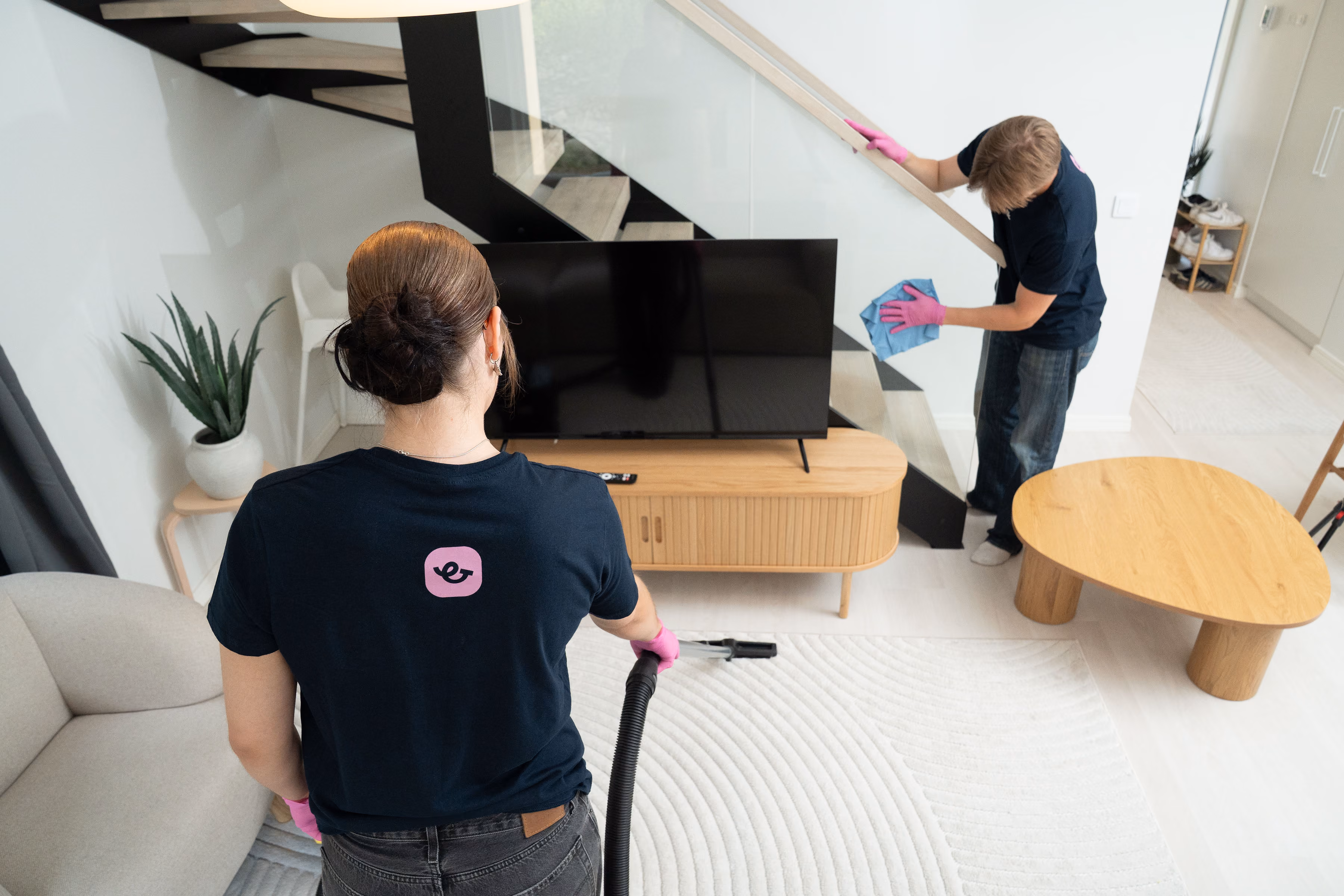 Two people cleaning a modern living room: one vacuuming a white rug, another wiping a handrail.