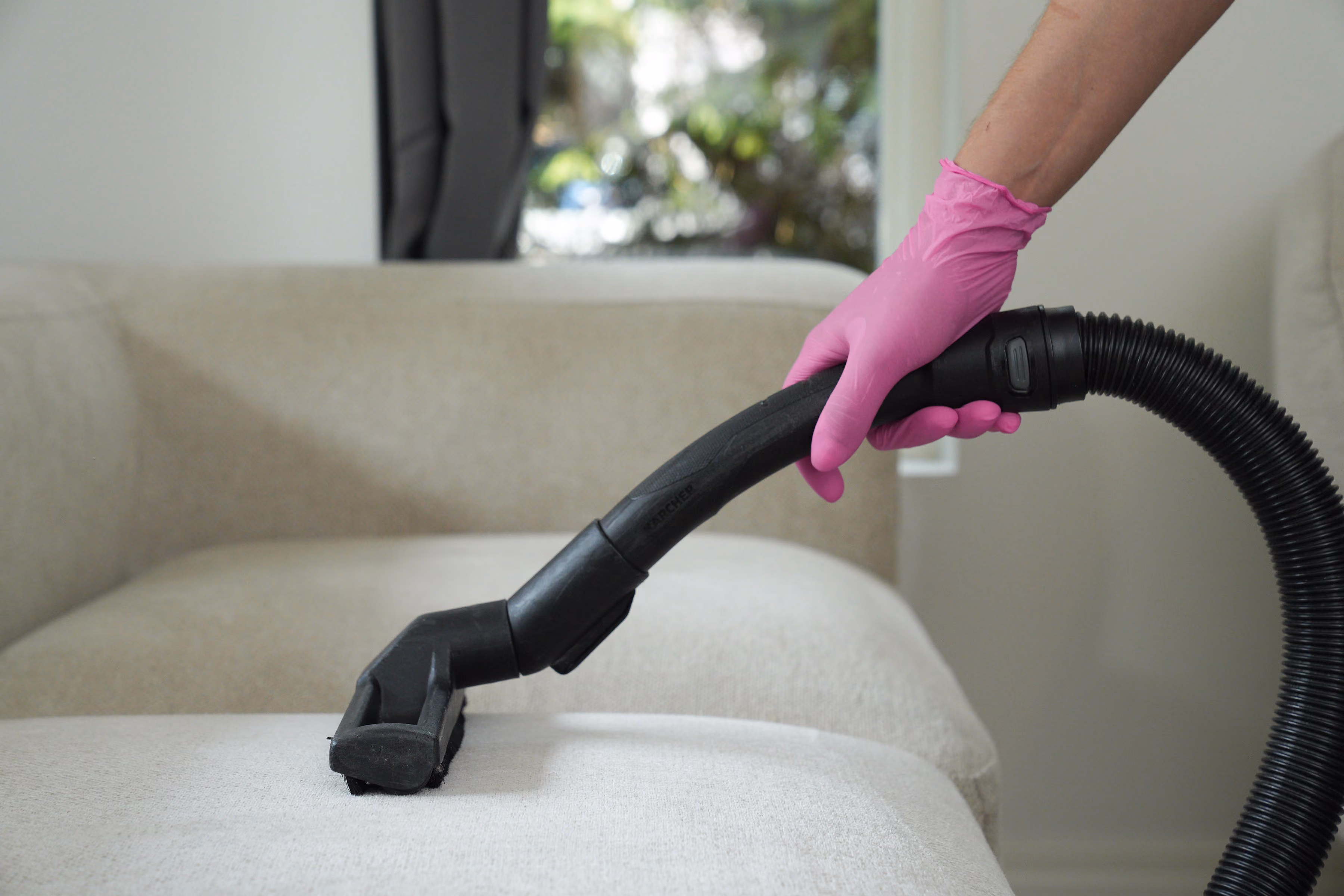 Hand with pink glove vacuuming a beige fabric sofa with a black hose attachment.