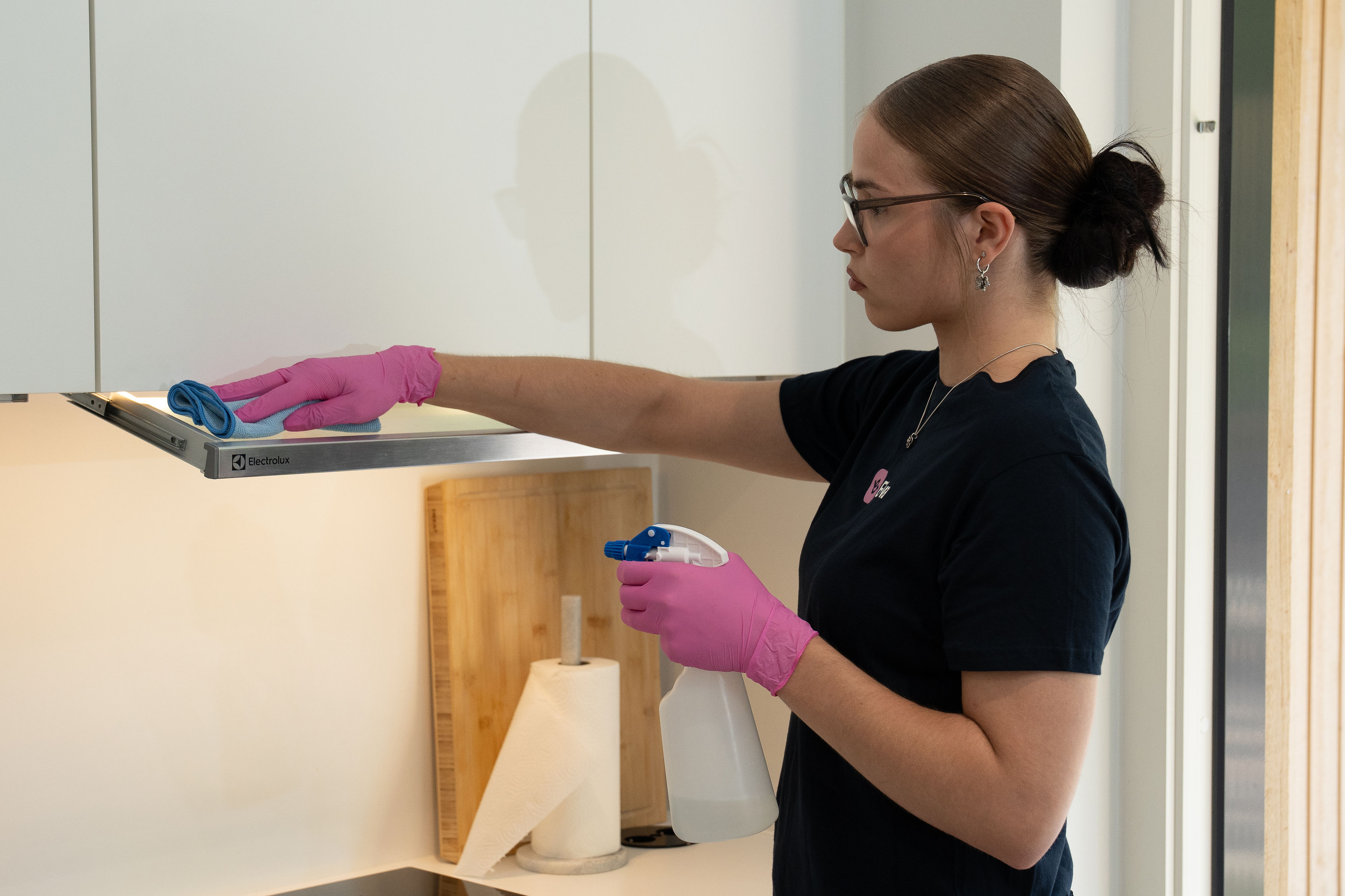 Woman wearing pink gloves cleaning kitchen hood with a blue cloth and spray bottle.