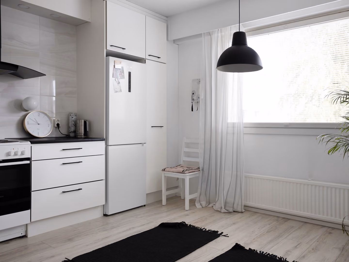 Minimalist kitchen with white cabinets, refrigerator, stove, black pendant light, and sheer window curtains.