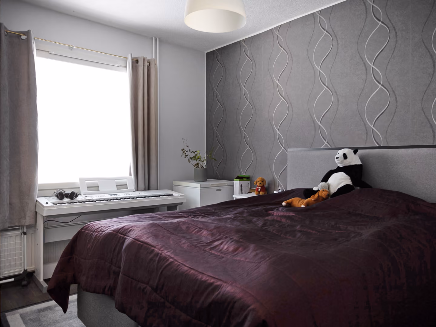 Bedroom with a purple bedspread, stuffed panda and toys on bed, keyboard by window, gray patterned wall.
