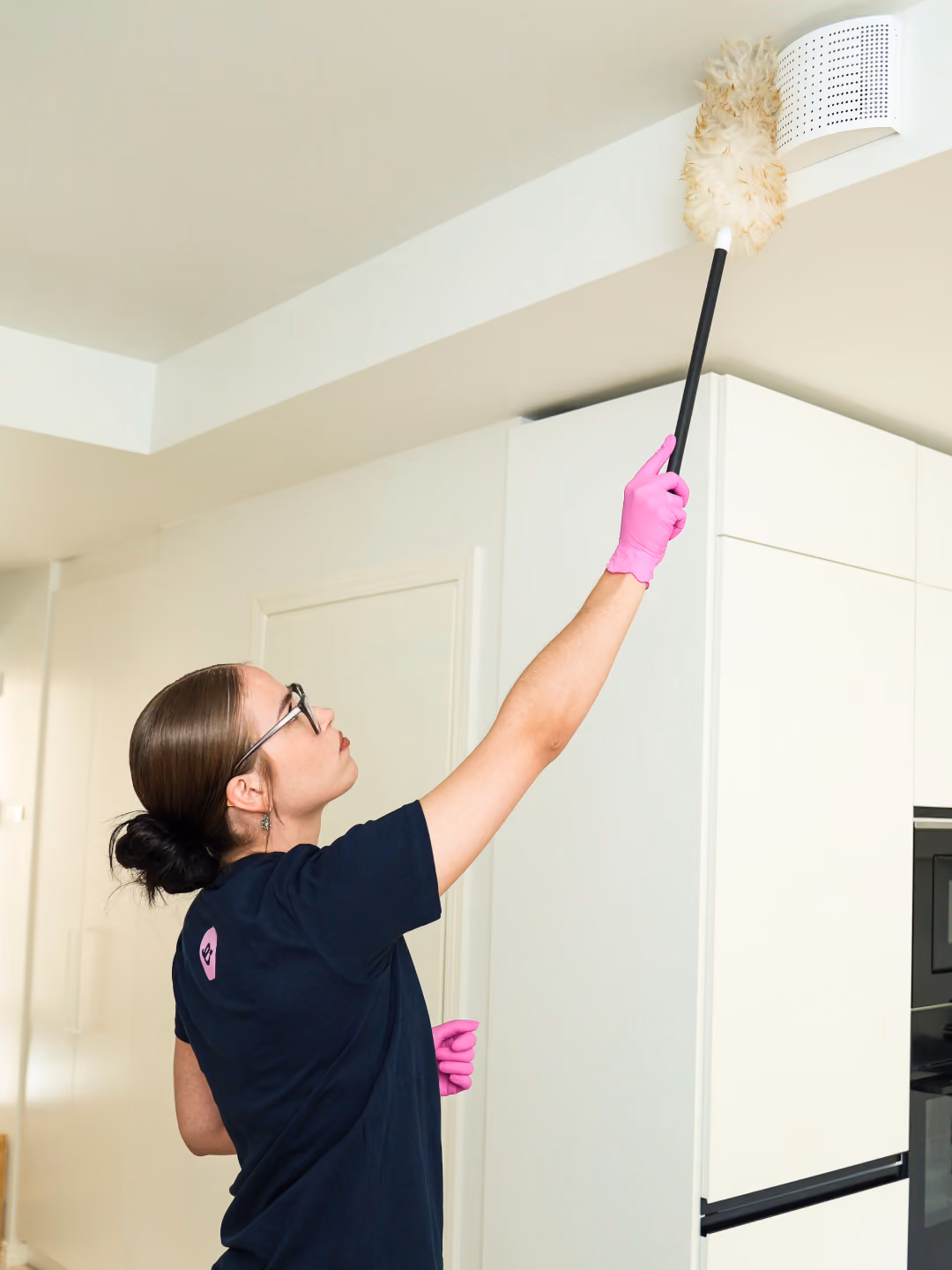 Woman wearing pink gloves dusting a ceiling vent with a long-handled duster in a kitchen.