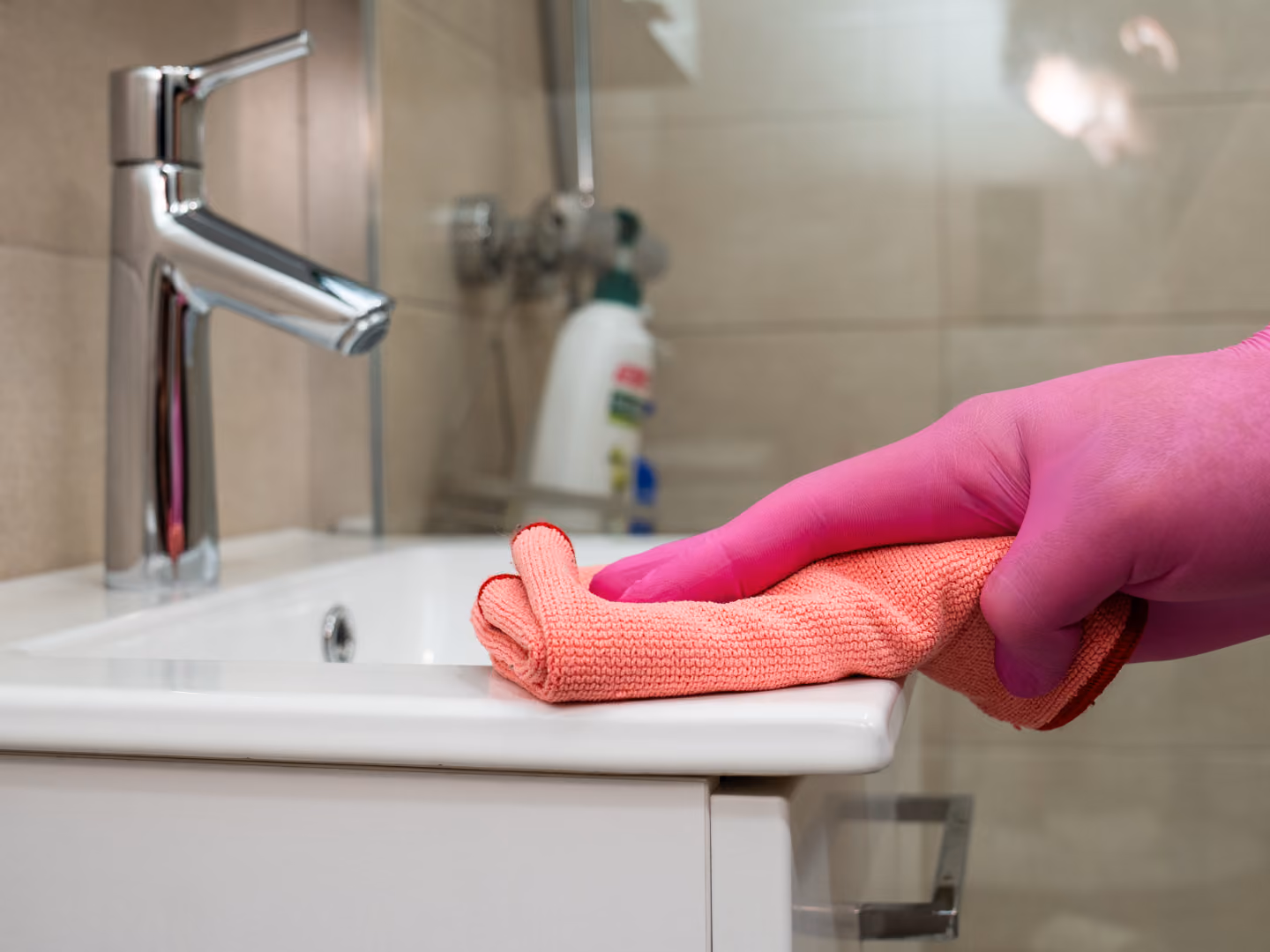 Hand in pink glove cleaning white bathroom sink edge with orange cloth.