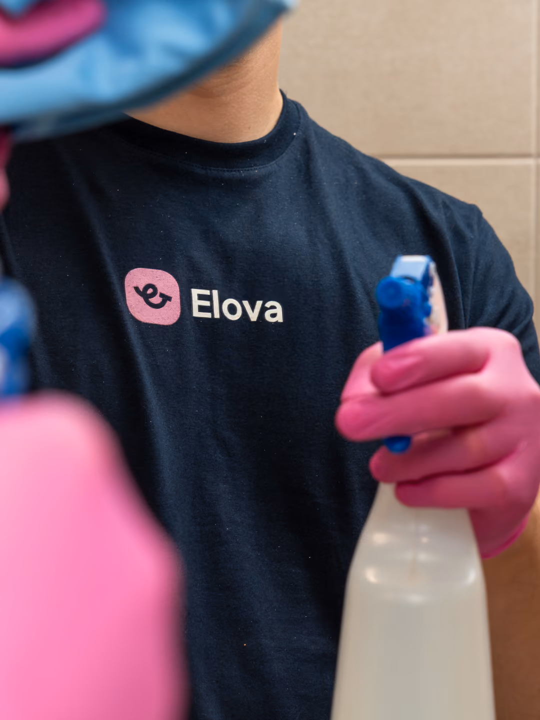 Person wearing a black Elova shirt and pink gloves holding a white spray bottle with blue nozzle.