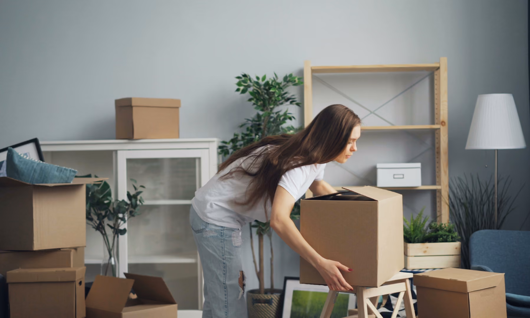 Woman in white shirt and jeans placing a cardboard box on a small wooden stool in a room with moving boxes.