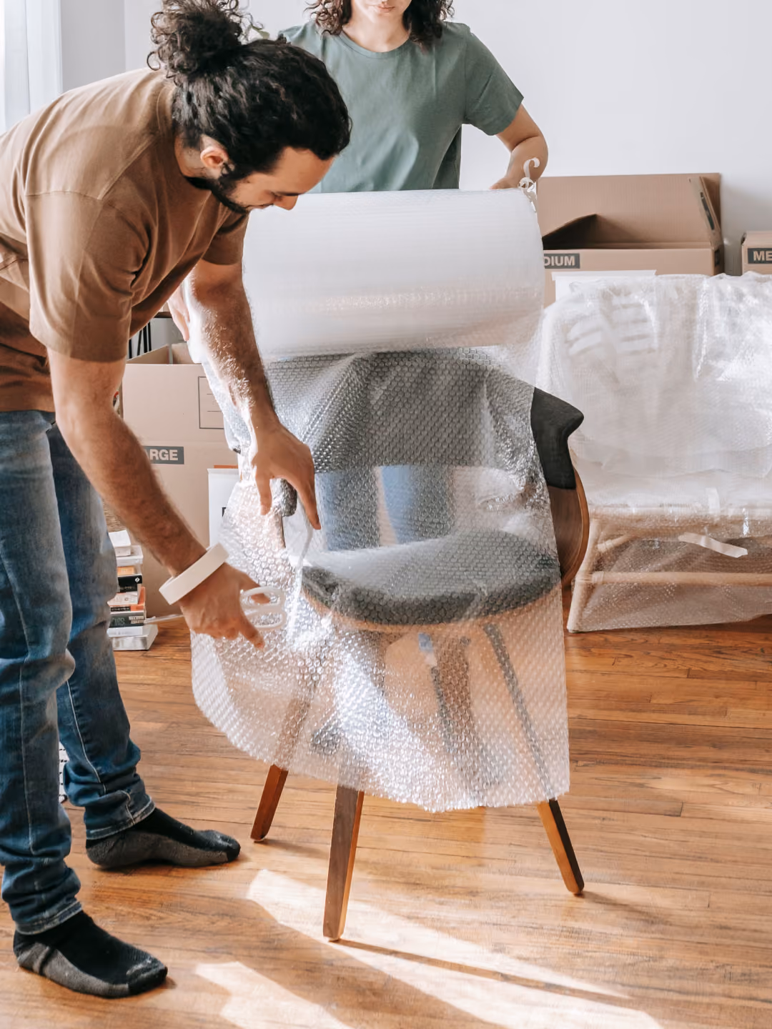 A man wrapping a chair with bubble wrap while a woman assists in a room with moving boxes.