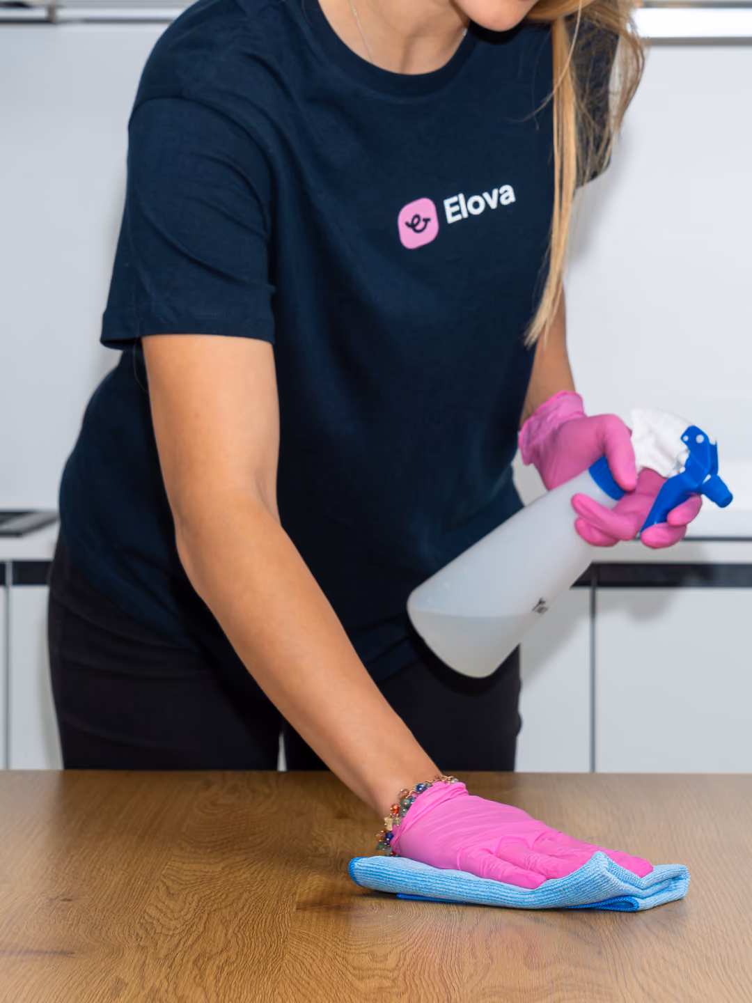 Person wearing pink gloves cleaning a wooden surface with a blue cloth and spray bottle.